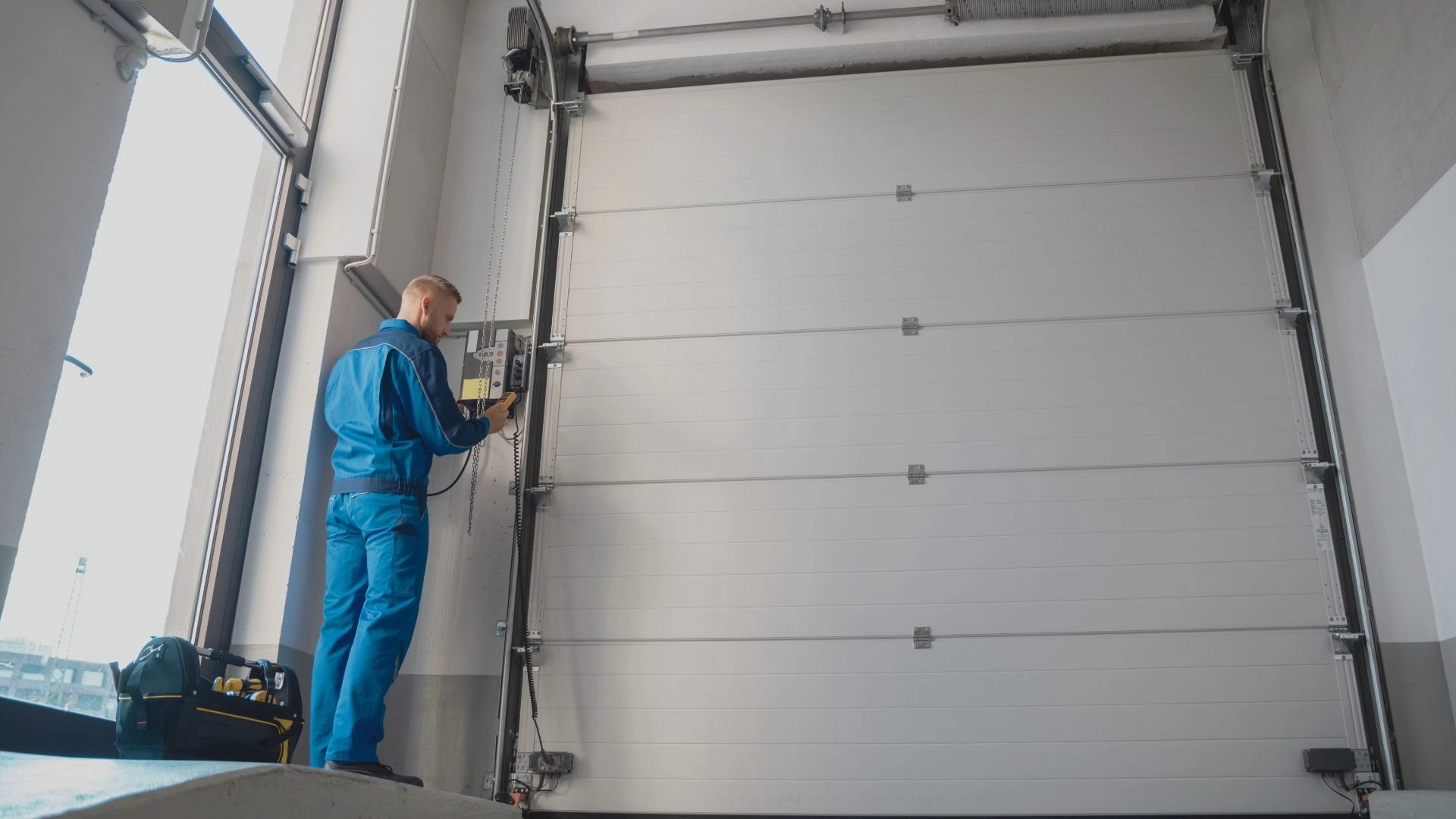 A technician in blue work attire operates a control panel beside a large, closed overhead garage door. A tool bag is positioned nearby.